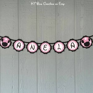 Op de afbeelding: Een zwart en roze banner met de naam "ANELA" gespeld in witte letters. Elke letter staat op een aparte zwarte cirkel met roze stippen. De uiteinden van de banner hebben een zwart en roze cartoon muizenkop.