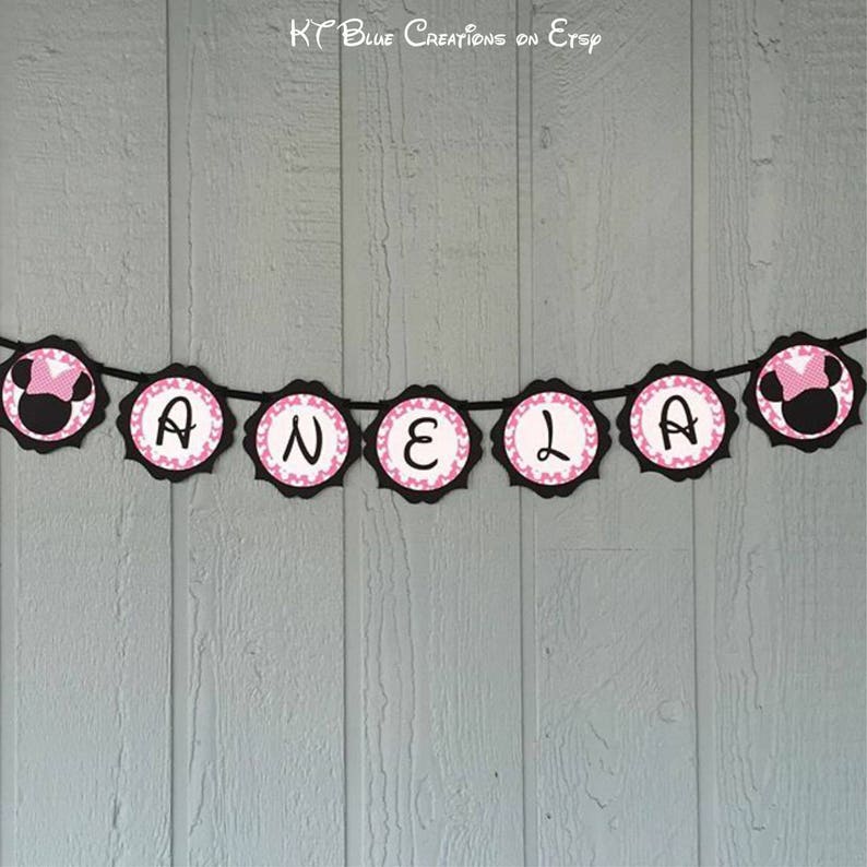 Op de afbeelding: Een zwart en roze banner met de naam "ANELA" gespeld in witte letters. Elke letter staat op een aparte zwarte cirkel met roze stippen. De uiteinden van de banner hebben een zwart en roze cartoon muizenkop.
