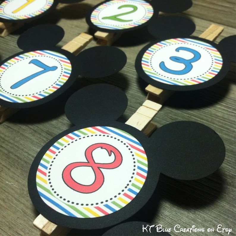 May include: Eight colorful Mickey Mouse head table number cards with a white background and black stripes. The numbers are in a circle with a white outline. The numbers are 1, 2, 3, and 8.
