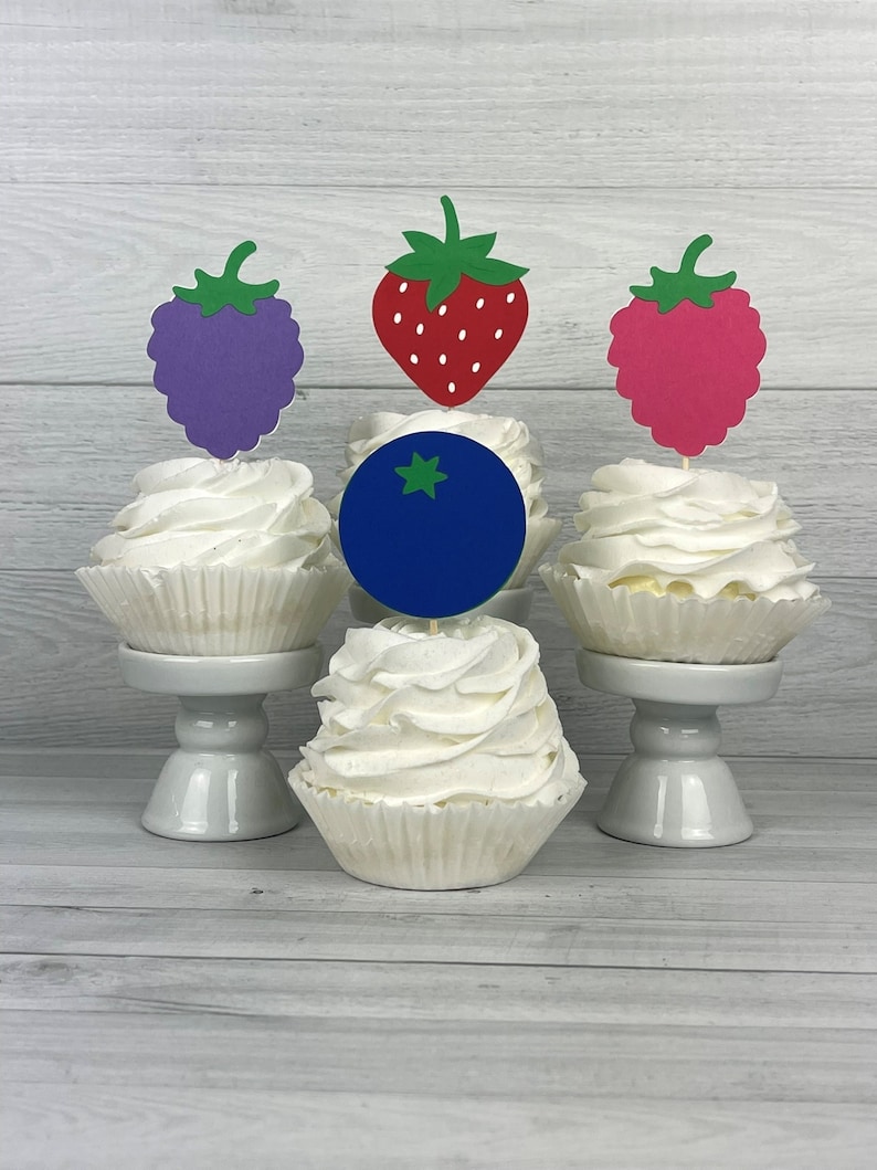Peut inclure: Quatre cupcakes d&eacute;cor&eacute;s de gla&ccedil;age blanc et de d&eacute;corations en forme de fruits. Les d&eacute;corations sont une fraise rouge, un raisin violet, une framboise rose et une myrtille bleue.