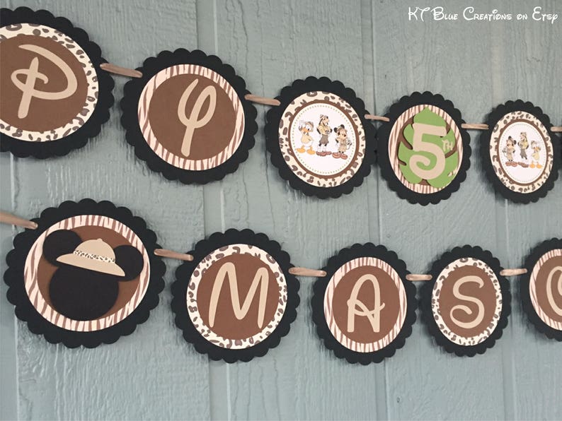 May include: A brown, black, and green banner with the letters "D", "P", "5", "M", "A", "S", and a cartoon image of Mickey Mouse wearing a safari hat. The banner is decorated with a zebra stripe pattern and leopard print.