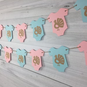 May include: A gender neutral baby shower banner with alternating blue and pink baby onesies with gold turkey silhouettes.