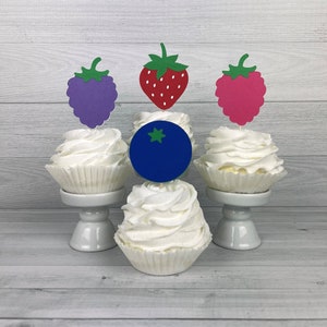 Peut inclure: Trois cupcakes d&eacute;cor&eacute;s de d&eacute;corations en forme de fruits. Les cupcakes sont blancs avec un tourbillon de gla&ccedil;age. Les d&eacute;corations sont une fraise rouge, un raisin violet et une framboise rose. Le cupcake du milieu a une d&eacute;coration de bleuet bleu.