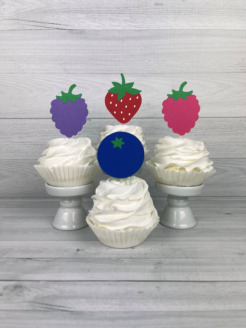 Peut inclure: Trois cupcakes d&eacute;cor&eacute;s de d&eacute;corations en forme de fruits. Les cupcakes sont blancs avec un tourbillon de gla&ccedil;age. Les d&eacute;corations sont une fraise rouge, un raisin violet et une framboise rose. Le cupcake du milieu a une d&eacute;coration de bleuet bleu.