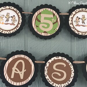 May include: A brown and black jungle-themed banner with a black and white zebra print, brown leopard print, and green leaves. The banner says "5th" and has images of Mickey Mouse, Donald Duck, and Goofy dressed in safari outfits.