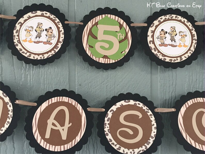 May include: A brown and black jungle-themed banner with a black and white zebra print, brown leopard print, and green leaves. The banner says "5th" and has images of Mickey Mouse, Donald Duck, and Goofy dressed in safari outfits.