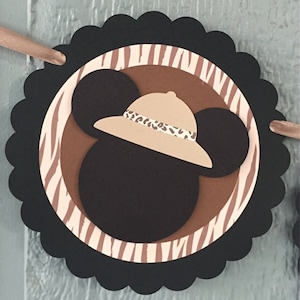 May include: Black and brown paper circle with a white and brown zebra stripe pattern. A brown Mickey Mouse head with a tan safari hat is in the center of the circle. The hat has a brown and tan leopard print band.
