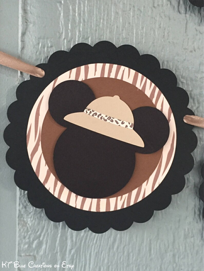 May include: Black and brown paper circle with a white and brown zebra stripe pattern. A brown Mickey Mouse head with a tan safari hat is in the center of the circle. The hat has a brown and tan leopard print band.
