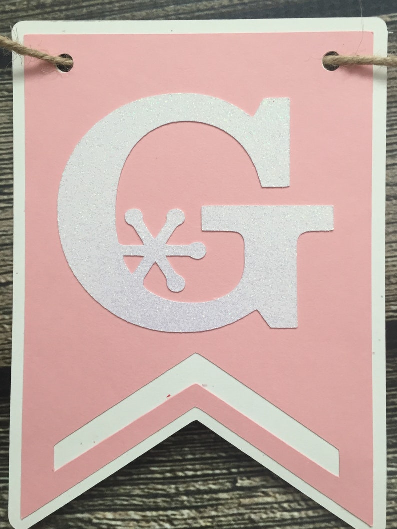 May include: Pink banner with a white glittery letter "G" and a snowflake-like design. The banner has a white border and a chevron design at the bottom. A twine string is attached at the top for hanging.