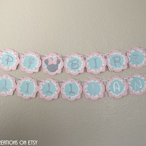 May include: A pink and blue banner with silver glitter letters spelling "Happy Birthday Lillian" with a silver glitter Minnie Mouse head on each end.