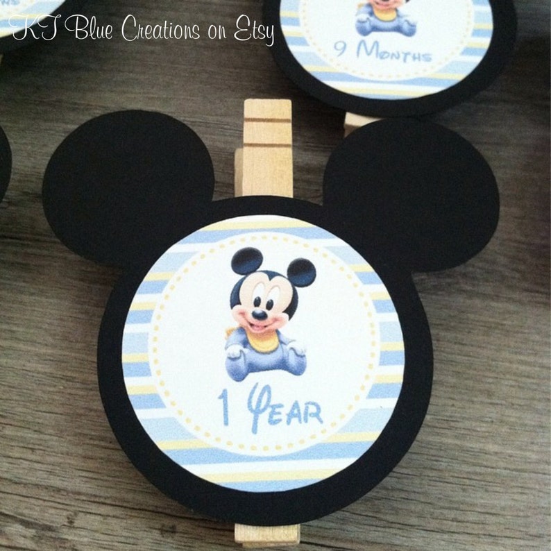 May include: Black and white Mickey Mouse character with blue clothing on a blue and yellow striped circle with the text "1 Year". The circle is attached to a black Mickey Mouse head cutout with a wooden clothespin.