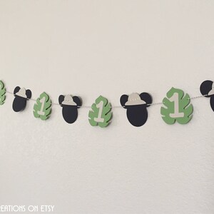 Puede incluir: Una pancarta de papel verde y blanca con orejas de Mickey Mouse y el n&uacute;mero uno en cada hoja. La pancarta est&aacute; enhebrada con una cuerda blanca. La pancarta es perfecta para una fiesta de primer cumplea&ntilde;os.