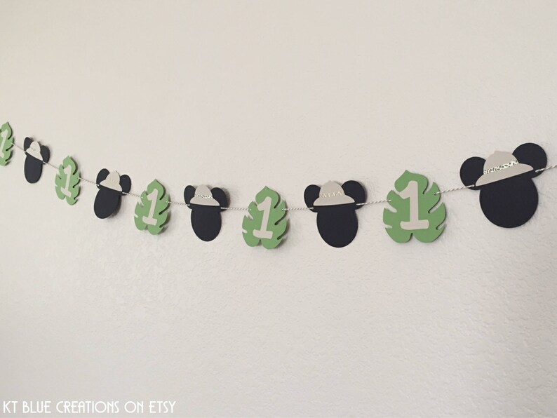 Puede incluir: Una pancarta de papel verde y blanca con orejas de Mickey Mouse y el n&uacute;mero uno en cada hoja. La pancarta est&aacute; enhebrada con una cuerda blanca. La pancarta es perfecta para una fiesta de primer cumplea&ntilde;os.