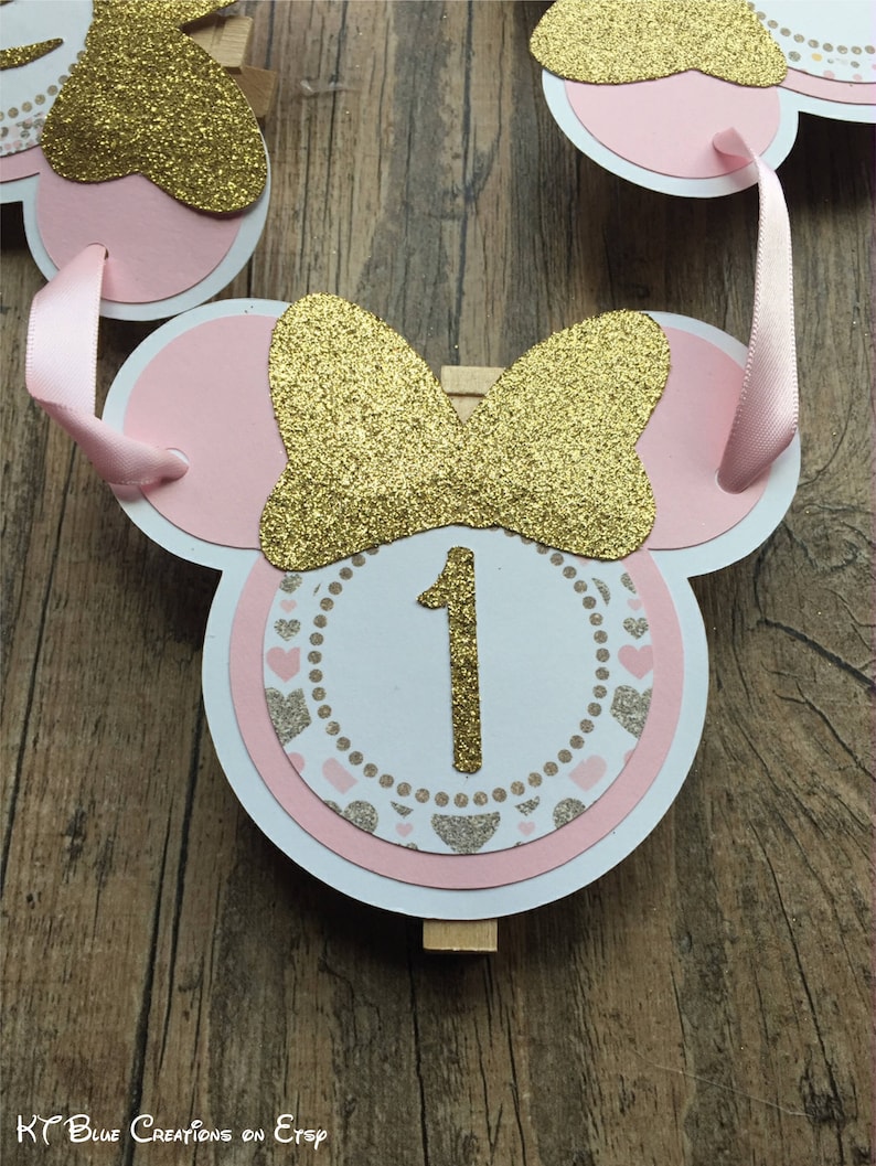 Op de afbeelding: Roze en gouden Minnie Mouse-themabanner met een witte cirkel met het nummer 1 in gouden glitter. De banner is aan elkaar geregen met roze lint en heeft een houten wasknijper.