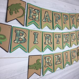 May include: A green, brown, and white camouflage patterned banner with the text "Happy 4th Birthday Reid" in brown letters. The banner has green and brown chevron accents and features cut-out images of a snake, lizard, and alligator.