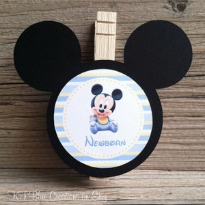 May include: Black and white Mickey Mouse head cutout with a blue and yellow striped circle in the center. The circle features a baby Mickey Mouse wearing a blue onesie and the word "NEWBORN".