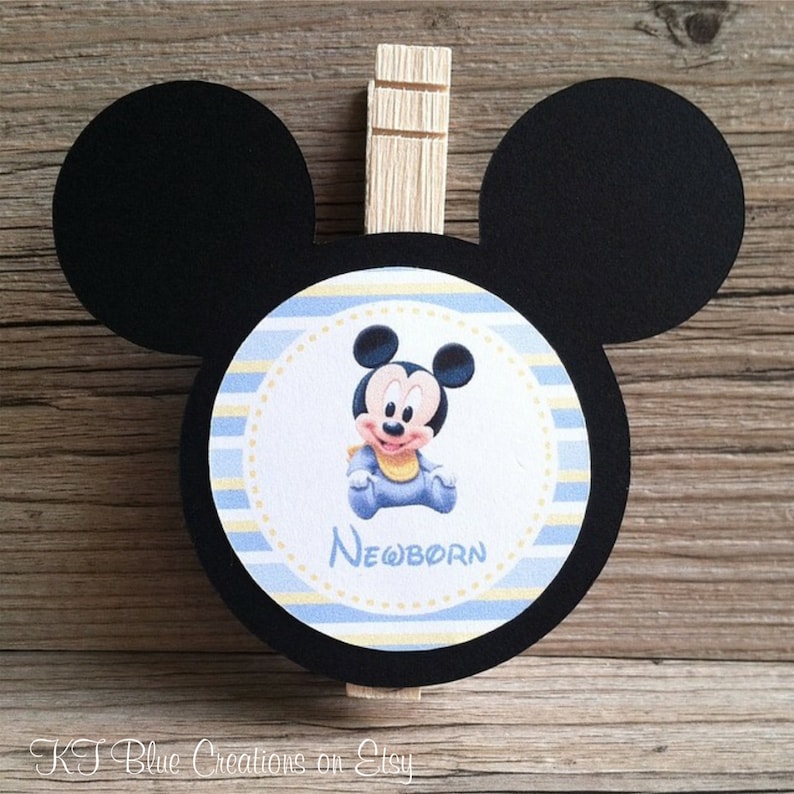 May include: Black and white Mickey Mouse head cutout with a blue and yellow striped circle in the center. The circle features a baby Mickey Mouse wearing a blue onesie and the word "NEWBORN".