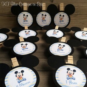 May include: Set of 12 black and white Mickey Mouse themed baby milestone cards with a blue and yellow striped background. The cards are numbered from newborn to 1 year and feature a cartoon image of Mickey Mouse in different poses. Each card has a wooden clothespin attached.