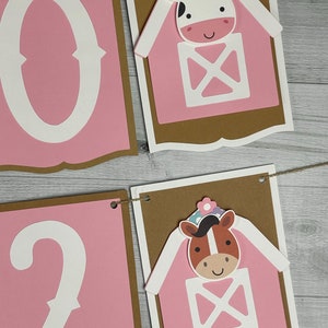 May include: Pink and brown banner with the numbers 0 and 2 in white. The banner has two pink barns with white doors and windows. A cow and a horse are peeking out of the barns. The barns have white flowers on top.