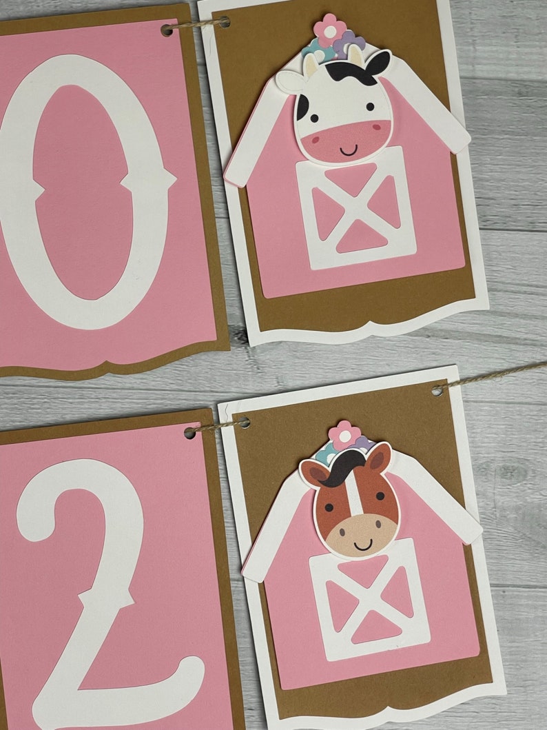 May include: Pink and brown banner with the numbers 0 and 2 in white. The banner has two pink barns with white doors and windows. A cow and a horse are peeking out of the barns. The barns have white flowers on top.