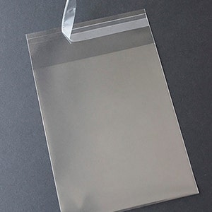 May include: A clear cellophane bag with a peel-off adhesive strip at the top. The bag is empty and slightly angled, with a grey background. The adhesive strip is partially peeled back, revealing the sticky surface.