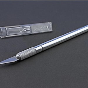 May include: A silver utility knife with a retractable blade and a clear plastic blade holder. The blade holder has the text "X-ACTO" printed on it.