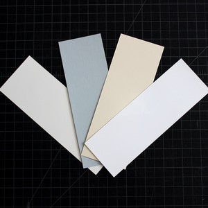 May include: Five rectangular swatches of paint chips in various shades of white, cream, and light blue. The swatches are arranged in a fan shape on a black grid.