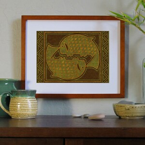 May include: A framed print of two stylized fish in a brown and green color scheme. The fish are facing each other and are surrounded by a geometric pattern.