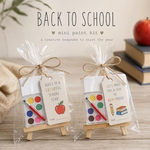 Op de afbeelding: Twee mini verfsets in doorzichtige plastic zakjes dichtgeknoopt met touw. Elke set bevat een klein canvas op een houten ezel, een verfpalet met zes kleuren en een penseel. De labels bevatten de teksten Here's to a colorful school year met een appel, en Let's make this a year to remember met een potlood en boeken.