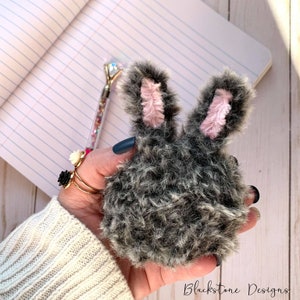 May include: A gray and black fuzzy bunny-shaped keychain with pink ears. The keychain is being held in a hand.