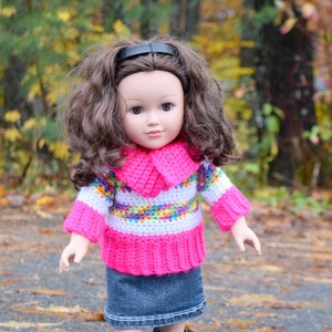 Könnte beinhalten: Eine Puppe trägt einen rosa-weißen gestreiften Häkelpullover mit einem bunten Muster, einen Jeansrock und schwarze Gummistiefel. Die Puppe hat braunes Haar und steht auf einem Weg mit heruntergefallenen Blättern.