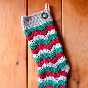 May include: A crocheted Christmas stocking with a white, red, and green striped pattern. The stocking has a small green wreath with red berries at the top.
