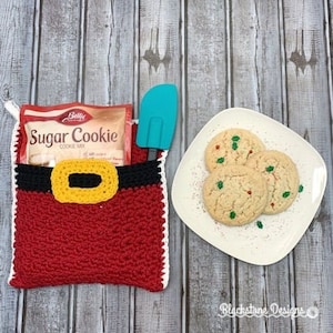May include: Red crocheted Santa Claus stocking with a black belt and yellow buckle. The stocking is filled with a box of Betty Crocker Sugar Cookie mix. A blue spatula and a plate of sugar cookies with green sprinkles are on a wooden surface.