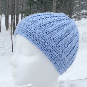 May include: A light blue crocheted beanie hat on a white mannequin head. The hat has a textured stitch pattern and is shown against a snowy background.