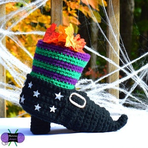 May include: A crocheted witch's boot with a green and purple striped top, black stars, and a gold buckle. The boot is filled with fall leaves and is sitting on a white surface.