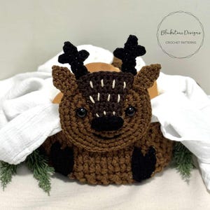 May include: A crocheted brown deer with black antlers, a black nose, and white accents. The deer has large black eyes and is sitting on a white surface. The image includes the text "Blackstone Designs Crochet Patterns."
