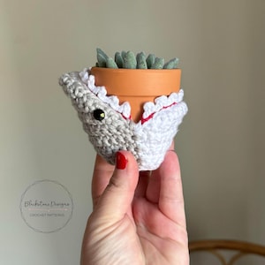 May include: A crocheted shark planter with a white and gray shark face and red teeth. The planter is holding a small succulent plant.