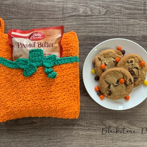 May include: Orange crochet pumpkin-shaped hot pad with a green crocheted bow and a packet of peanut butter inside. A white plate with three chocolate chip cookies and orange and yellow sweets is on a wooden surface.