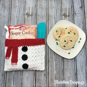Puede incluir: Una bolsa de regalo de crochet blanco con forma de muñeco de nieve con una bufanda roja y botones negros. La bolsa contiene una caja de mezcla para galletas de azúcar Betty Crocker y una espátula azul. Un plato blanco con tres galletas de azúcar decoradas con chispas rojas y verdes se encuentra sobre una superficie de madera.