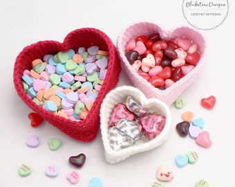 May include: Three crocheted heart-shaped baskets filled with candy. The largest basket is red and filled with pastel heart-shaped candies. The middle basket is pink and filled with red, pink, and brown jelly beans. The smallest basket is white and filled with chocolate candies. There are also scattered heart-shaped candies on the white background.