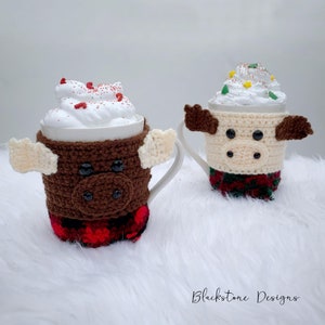 May include: Two white mugs with whipped cream and decorative sprinkles, each adorned with a crocheted animal cozy. One is a brown moose with cream-colored antlers and a red plaid base. The other is a cream-colored cow with brown ears and a green and red base.