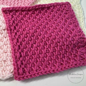 May include: A square, handmade crochet item in a vibrant fuchsia color. The textured stitch pattern creates a raised, geometric design. The item is part of a collection of crochet pieces, with a light pink and white item visible in the background.