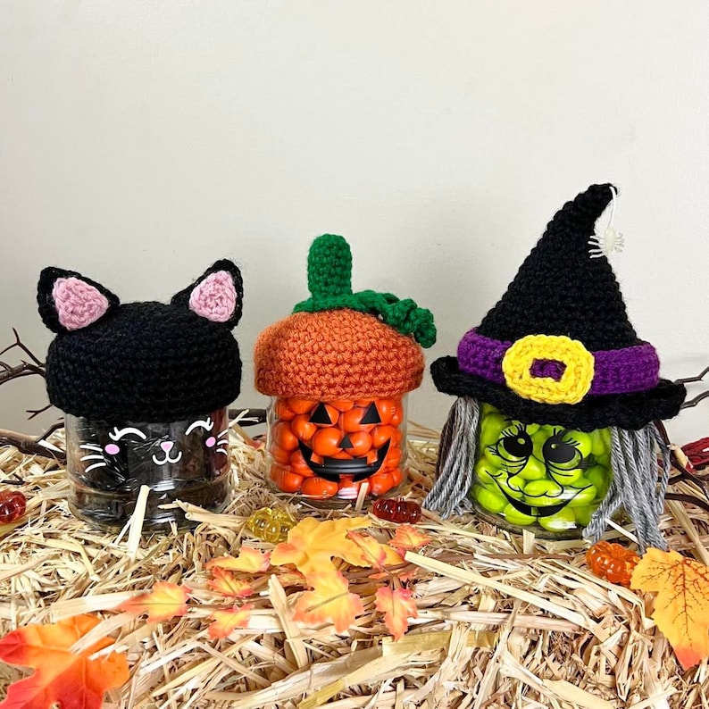 K&ouml;nnte beinhalten: Drei Halloween-Gl&auml;ser mit geh&auml;kelten Deckeln. Die Gl&auml;ser sind mit S&uuml;&szlig;igkeiten gef&uuml;llt und haben bemalte Gesichter. Ein Glas hat einen schwarzen Katzendeckel, eines einen K&uuml;rbisdeckel und eines einen Hexenhutdeckel.