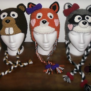 May include: Three crocheted hats with animal faces. The hats are brown, orange, and gray with black, white, and orange braids. The hats are on white mannequins with the braids hanging down.