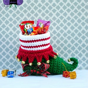 May include: A green, red and white crocheted elf shoe with jingle bells, filled with wrapped sweets and small gifts. The shoe is sitting on a white surface with a grey patterned background.