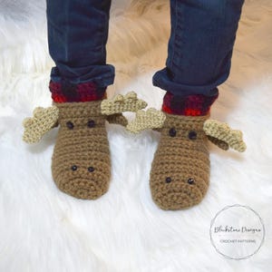 May include: A pair of brown crocheted moose slippers with black eyes and red and black plaid cuffs. The slippers are worn by a person wearing blue jeans.