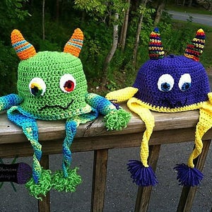 May include: Two crocheted monster hats with long tails. One hat is green with orange and blue accents, the other is purple with yellow accents.