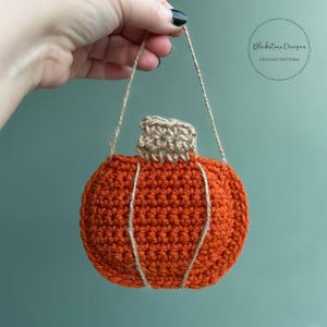 Puede incluir: Un adorno de calabaza de ganchillo con un tallo marrón y una cuerda de color beige para colgar. La calabaza es de color naranja y tiene una superficie texturizada.