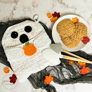 Puede incluir: Un agarrador de ganchillo blanco con forma de fantasma con ojos y boca negros, sosteniendo una calabaza naranja. Un plato de galletas y dos espátulas grises y con mango de madera están cerca. La escena está decorada con hojas de otoño.