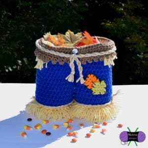 Peut inclure: Un récipient en crochet bleu fait à la main ressemblant à un pantalon d'épouvantail, rempli de feuilles d'automne. Le récipient a un bord marron et est décoré d'un bouton et de liens en fil. Des franges en fil jaune et des bonbons complètent le thème de l'automne.